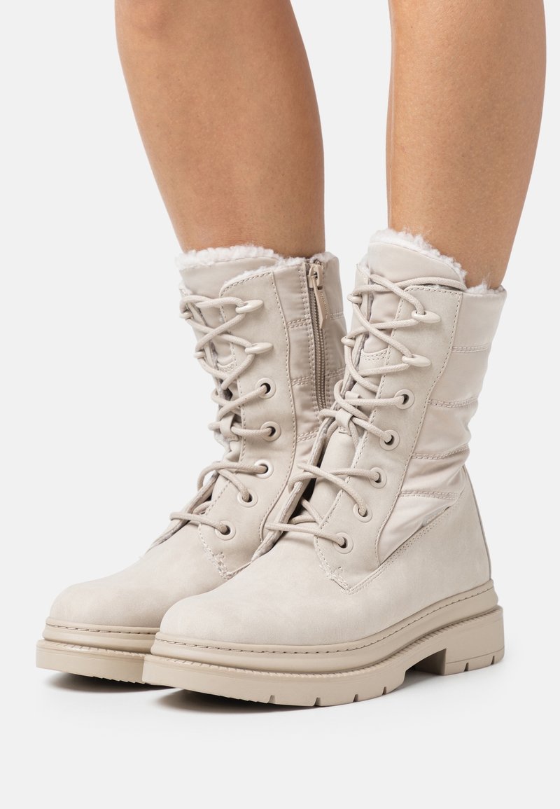 Bottes d'hiver lacées beige clair avec fermetures éclair latérales, semelles épaisses et doublure en fausse fourrure portées sur les jambes inférieures sur fond blanc.