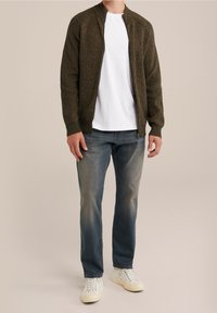 Oliivinvihreä vetoketjullinen neule, jossa on kudottu rakenne, yhdistetty valkoiseen t-paitaan ja haalistuneisiin sinisiin farkkuihin, ja viimeistelty valkoisilla lenkkareilla.