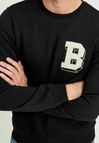 Crna dukserica s okruglim izrezom, koja na prsima ima teksturirano bijelo slovo "B" u obliku bloka. Rebrasti rukavi i donji rub dodaju detalj.