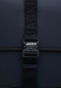 Navyblå taske med en glat overflade, udstyret med en sort rem prydet med "JACK1T" og en sort plastikspænde til sikker fastgørelse.