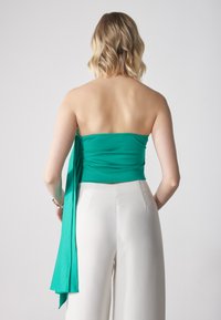 Grön strapless topp med en slät textur och en sidoknytning, tillsammans med vita högmidjade byxor som visar en figurnära silhuett.