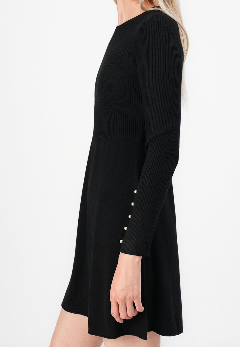 Femme portant une robe noire côtelée à manches longues avec des boutons en perles sur le poignet, vue de profil sur fond blanc.