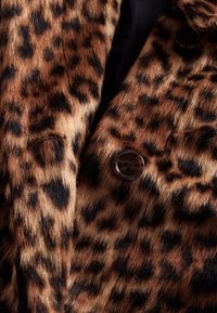 Manteau en fausse fourrure à imprimé léopard avec une texture douce, présentant des taches marron et noires, une fermeture à boutons et des détails de couture visibles.