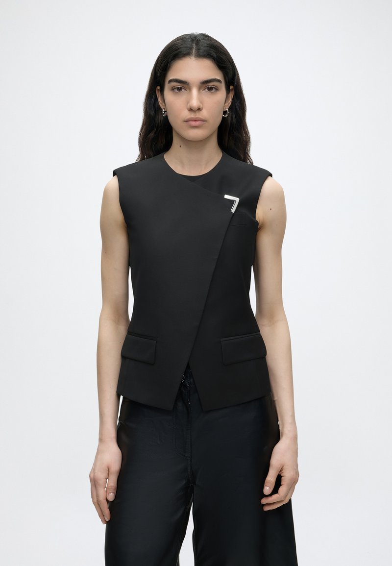 Mujer con cabello largo y oscuro lleva un chaleco negro sin mangas con frente asimétrico y alfiler plateado, combinado con pantalones negros sueltos, fondo neutro.