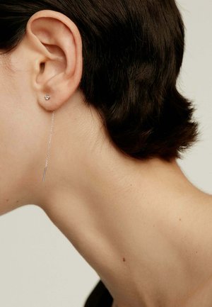 Los pendientes de plata tienen un pequeño stud y una cadena delgada con una barra recta y minimalista colgando debajo del lóbulo de la oreja.