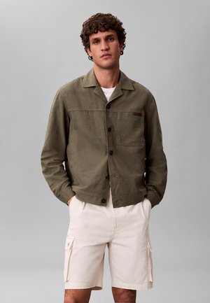 Mężczyzna w oliwkowo-zielonej zapinanej na guziki kurtce na białej koszuli i jasnobeżowych spodenkach typu cargo, stojący z rękami w kieszeniach na szarym tle.