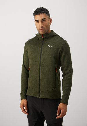 Άνδρας που φοράει πράσινο φούτερ με φερμουάρ και πορτοκαλί τράβηγμα και μαύρο παντελόνι, στέκεται μπροστά σε απλό ανοιχτόφωτο φόντο.