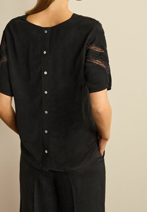 Blusa negra de manga corta con detalles de encaje transparente en los hombros, escote redondeado y una fila de seis botones en la parte posterior. Textura suave.