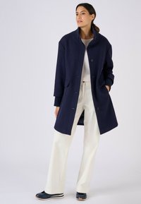 Cappotto in misto lana color blu navy con collo alto, chiusura con bottoni e tasche laterali. Indossato sopra un maglione leggero e pantaloni larghi e chiari.
