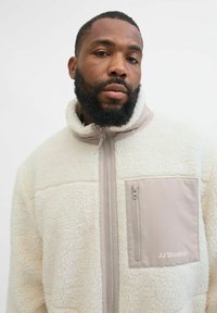 Giacca in pile cremoso con collo alto, zip frontale e tasca sul petto beige con chiusura a zip e logo "JJ Studios".