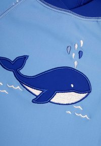 Tissu bleu clair arborant une baleine brodée en bleu foncé et blanc, avec des gouttes d'eau et des vagues en accents de fil blanc.