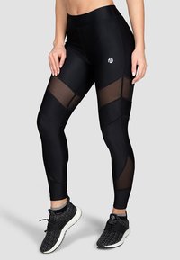 Černé sportovní legíny s síťovinovými panely, vysokým pasem a geometrickými designovými akcenty. Logo na přední straně. Spárované s černými sportovními botami.