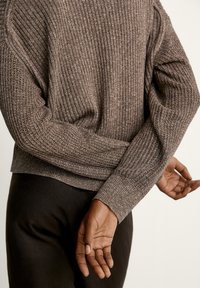 Brauner gerippter Pullover mit strukturiertem Strickmuster und enganliegenden Bündchen, kombiniert mit dunkler, hoch taillierter Hose. Rückansicht zeigt die Positionierung der Arme.