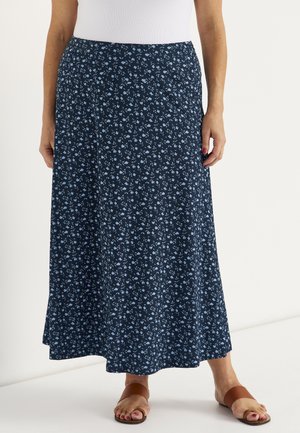 Vrouw draagt een marineblauwe maxirok met een klein lichtblauw bloemenpatroon, een wit topje en bruine platte sandalen.