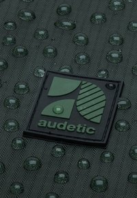 Superficie verde testurizzata con gocce d'acqua circolari. Un logo rettangolare nero è ben visibile, con un design geometrico e il nome del marchio.