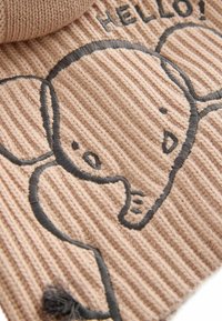 Gestrickter beiger Stoff mit horizontalen Rippen, featuring einen schwarzen gestickten Elefanten mit dem Text "HALLO!" darüber. Weiche Textur und abgerundete Formen.