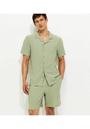 Mężczyzna ubrany w jasnozieloną koszulę z krótkim rękawem zapinaną na guziki oraz pasujące szorty, stoi z jedną ręką w kieszeni na białym tle.