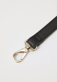 Musta teksturoitu hihna, jossa on kultainen metallinen kääntöklipsi, joka on varustettu sileällä pinnalla ja vähäisellä ommella. Suunniteltu kiinnittämään esineet turvallisesti.