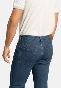 Vaqueros de mezclilla azul oscuro con un diseño ajustado, que cuentan con dos bolsillos traseros y costuras en contraste. Llevados con una camisa deportiva de color claro.