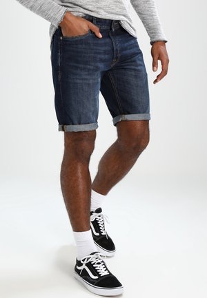 Mand iført opkrogede mørke denimshorts, grå langærmet skjorte, hvide ankelsokker og sorte lave sneakers står med den ene hånd i lommen.