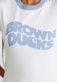 Hvit bomull t-skjorte med lyseblå ribbkrage. Fronten har stor, fet tekst "BROWN DUCKS" i lyseblå. Jevn tekstur.