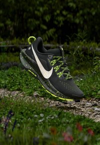 Schwarzer Sportschuh mit weißem Swoosh, grünen Akzenten und gesprenkelter Sohle. Verfügt über ein Mesh-Obermaterial, strukturierte Schnürung und eine gepolsterte Zwischensohle.