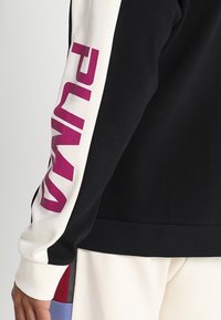 Sweatshirt noir avec des manches blanches, présentant un grand logo "PUMA" violet. Tissu texturé, poignets côtelés et un design classique à col rond.