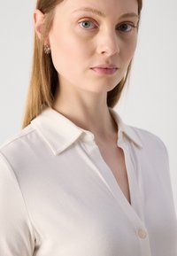 Krém színű, galléros gombos ing; puha anyagú textúra; elülső gombolása és finom fényessége van; letisztult, egyszerű dizájn.