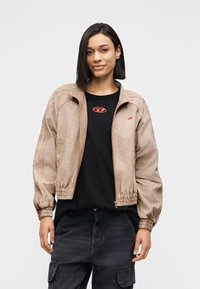 Ανοιχτό καφέ ζακέτα με φερμουάρ, ελαστικές μανσέτες και τελείωμα, με ομαλή υφή και διακριτική λάμψη, φορεμένη πάνω από μαύρο t-shirt με κόκκινο λογότυπο.