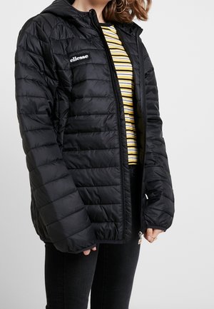 Femme portant une veste matelassée noire ellesse par-dessus une chemise rayée jaune et noire et un pantalon noir, debout devant un fond uni.