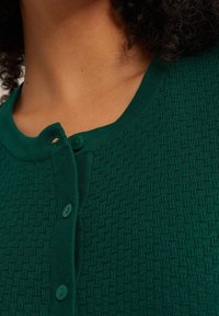 Grön cardigan med ett strukturerat mönster, rund halsringning och gröna knappar längs framsidan. Tyget verkar mjukt och stickat.