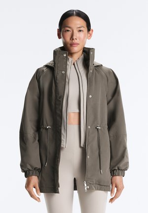 Žena v taupe bunde s vysokým golierom na zips cez svetlobéžový krátky top a ladiace legíny s vysokým pásom, stojaca na bielom pozadí.