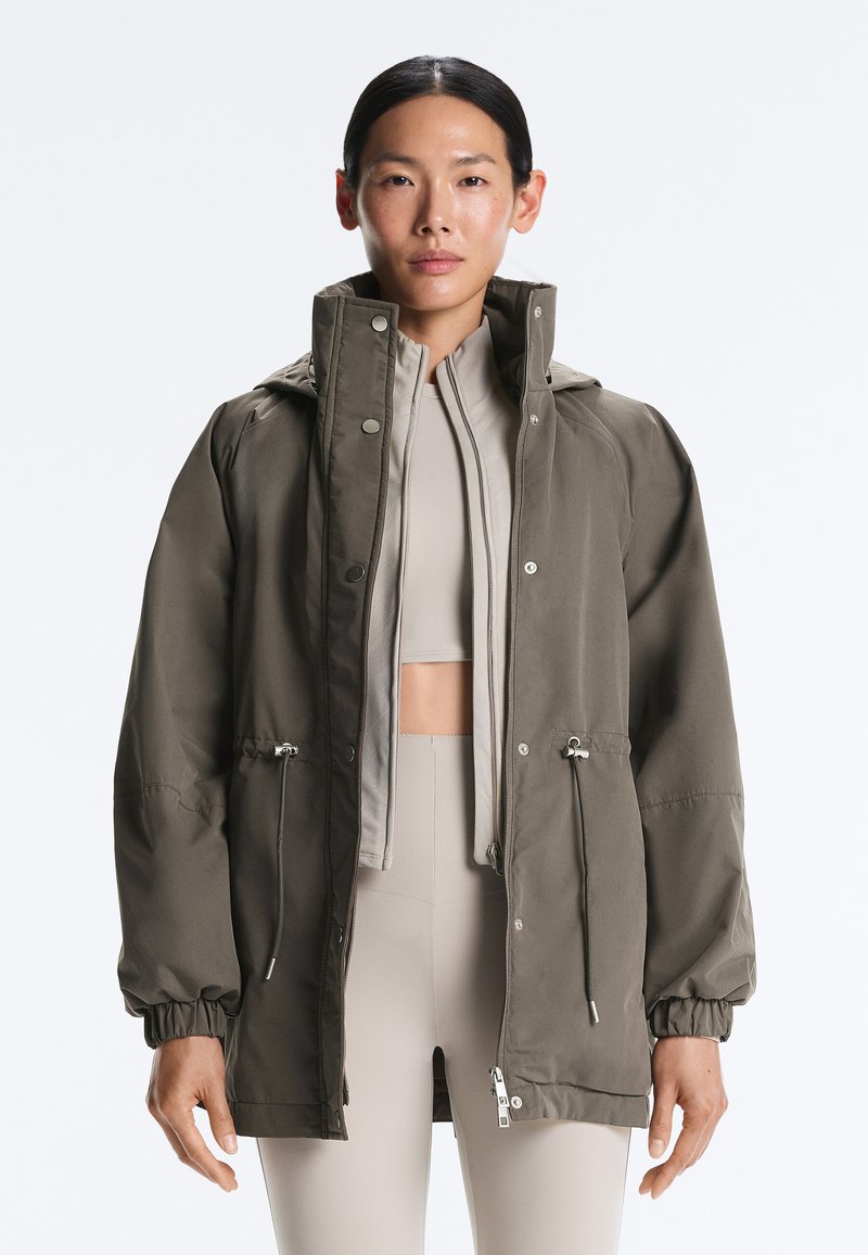 Kobieta ubrana w taupe kurtkę z wysokim kołnierzem na zamek błyskawiczny, na jasnobeżowym krótkim topie i pasujących legginsach z wysokim stanem, stojąca na białym tle.