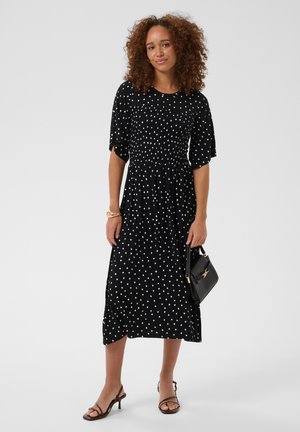 Femme aux cheveux bouclés portant une robe midi noire à pois blancs, tenant un sac à main noir et portant des talons noirs à lanières.