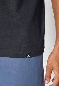 T-shirt en coton noir à manches courtes, avec une petite étiquette blanche du logo Adidas au bas, associé à un short de sport bleu.