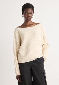 Off-white neulepaita, jossa on näyttävä, leveä kaula-aukko ja ribbikuvio. Ominaisuuksia ovat roikkuvat olkapäät ja rento istuvuus, yhdistetty mustiin cargo-housuihin.
