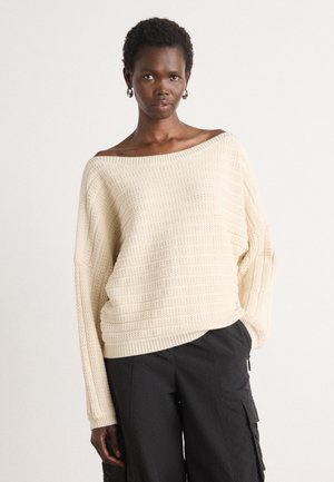 Off-white knit sweater with a loose, wide neckline and ribbed texture. Features dropped shoulders and a relaxed fit, paired with black cargo pants.
