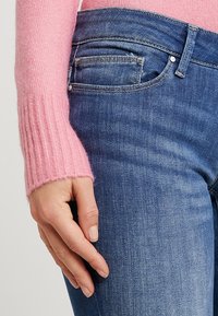 Handen vilar vid sidan, iklädd en rosa ribbad långärmad tröja och blå denimjeans med synlig främre ficka och bältesstropp.