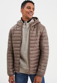 Braune Pufferjacke mit Kapuze, mit einem gesteppten Design, Frontreißverschluss und geripptem Kragen, über einem strukturierten hellen Pullover und blauen Jeans.