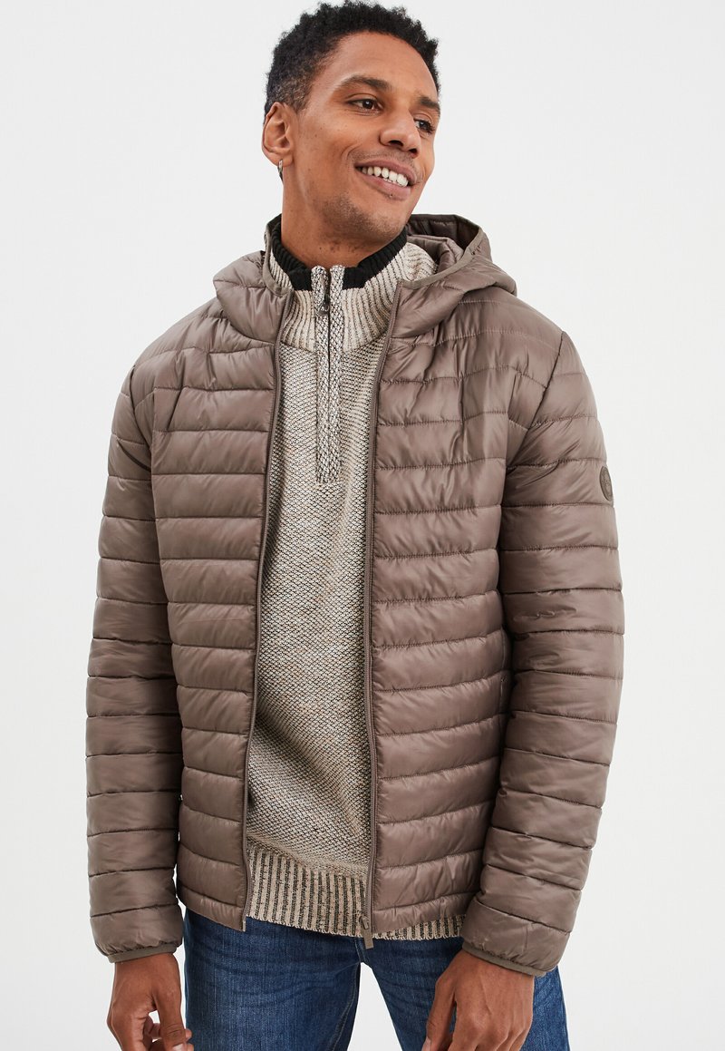 Braune Pufferjacke mit Kapuze, mit einem gesteppten Design, Frontreißverschluss und geripptem Kragen, über einem strukturierten hellen Pullover und blauen Jeans.