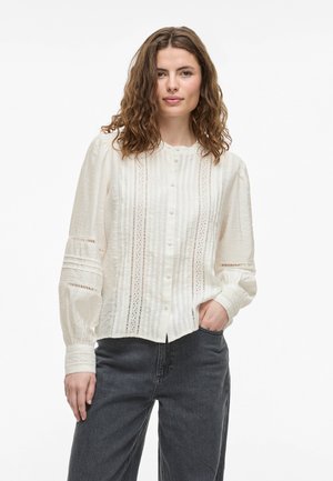 White button-up blouse with vertical stripes and eyelet detailing, featuring long sleeves and a rounded neckline. Worn with dark gray pants.
