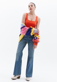 Top de tirantes ajustado naranja, cárdigan de punto multicolor con diseño floral, vaqueros de cintura alta azules y botas de tacón blancas por el tobillo.