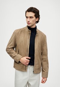 Νεαρός άνδρας με σκούρα, σγουρά μαλλιά, φορώντας μπεζ σουέτ τζάκετ πάνω από μαύρο ζιβάγκο και ανοιχτόχρωμο παντελόνι, κοιτάζοντας στο πλάι με το χέρι κοντά στο φερμουάρ.