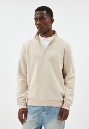 Pull-over beige clair à fermeture éclair au quart, en matériau doux, comportant un ourlet et des poignets côtelés, superposé sur une chemise blanche, avec une texture lisse.