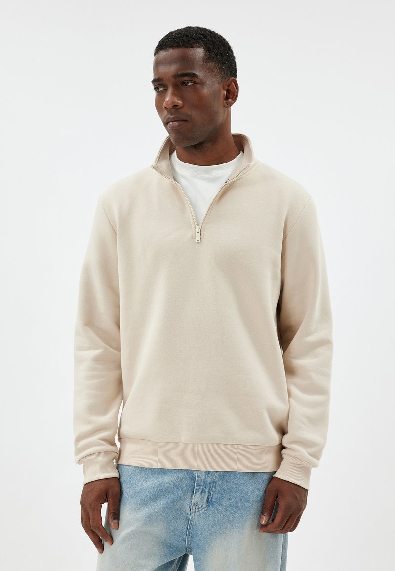 Sudadera de cuello alto color beige claro, hecha de un material suave, con dobladillo y puños acanalados, superpuesta sobre una camisa blanca, con una textura lisa.