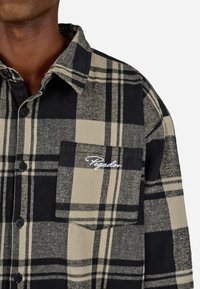 Fekete és bézs kockás flaneling, gombos elülső résszel, gallérral, mellzsebbel és hímzett logóval a zseben.