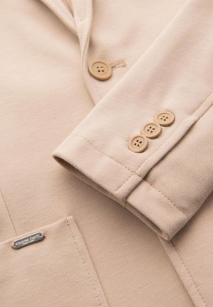 Manica di blazer beige con tre bottoni beige e tasca frontale con una piccola etichetta del marchio in argento.