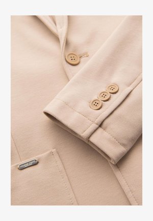 Manica di blazer beige con tre bottoni beige e tasca frontale con una piccola etichetta del marchio in argento.
