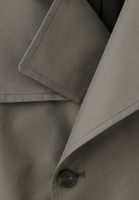 Gros plan du col et du revers d'un trench-coat vert olive avec un bouton noir visible et des détails de coutures piquées.