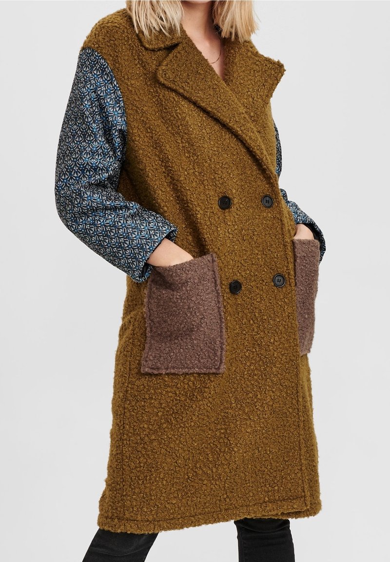 Femme portant un manteau marron texturé à double boutonnage avec de grandes poches mauves sur une chemise à manches longues à motif, les mains dans les poches.